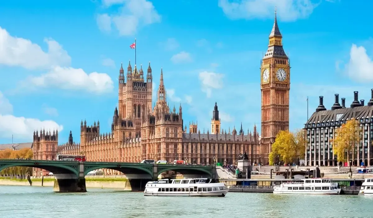 A scenic daytime view of the Houses of Parliament and Big Ben in London, with boats cruising on the River Thames and the UK flag waving atop the building.