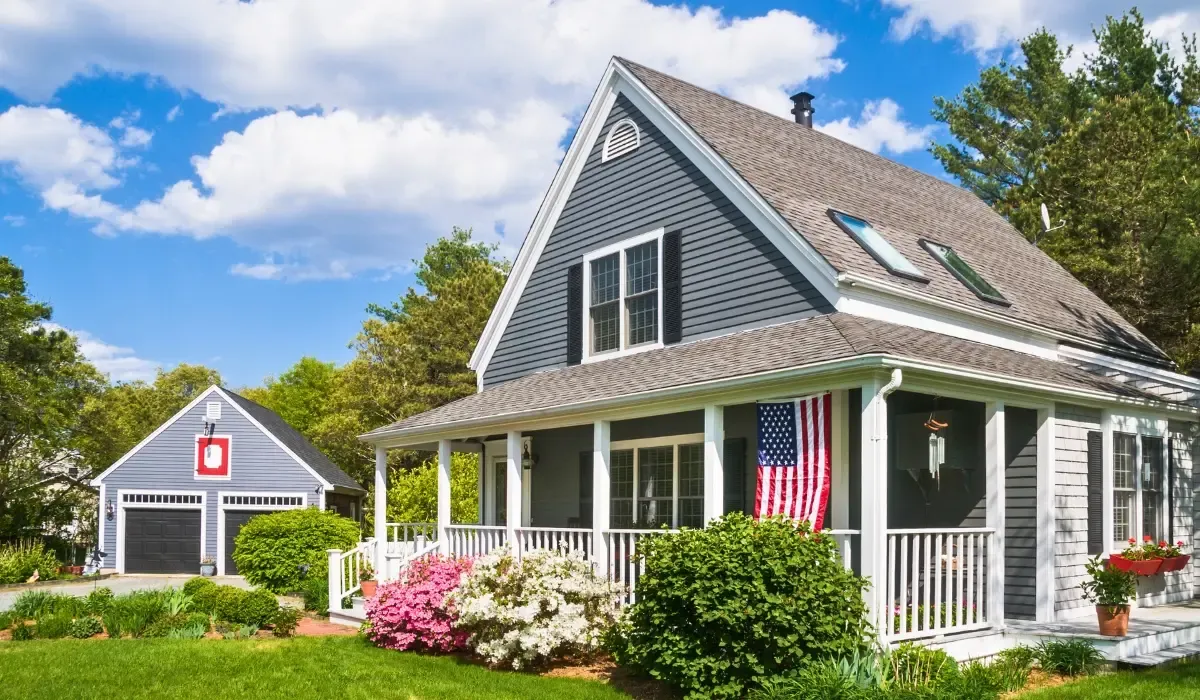 A modern American suburban house with a flag on the porch, surrounded by blooming flowers and greenery under a sunny blue sky.