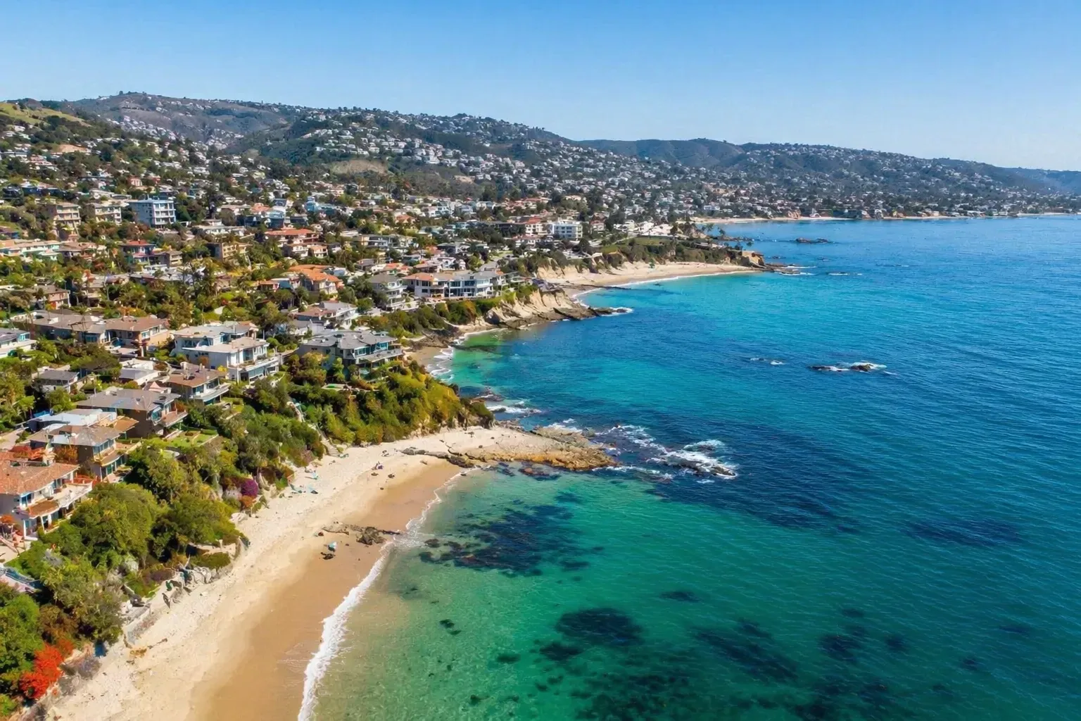 Aerial view of a tropical coastal neighbourhood with luxury homes, sandy beaches, turquoise ocean water, and rocky shoreline. Aerial view of a scenic coastal community with hillside homes, sandy beach, clear turquoise water, and a sweeping ocean shoreline.