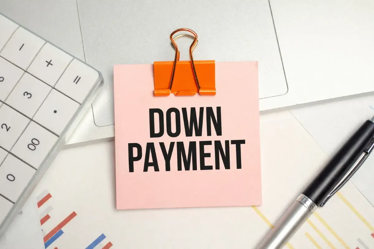 Pink sticky note reading down payment clipped with orange binder clip beside calculator, laptop, charts, and pen on office desk.