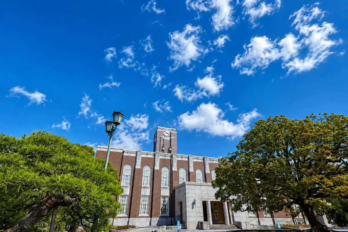 Kyoto University’s historic clock tower building framed by lush green trees under a bright blue sky with scattered white clouds.