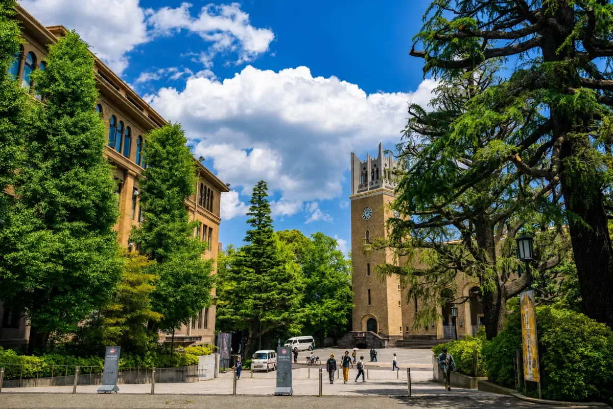 Waseda University campus entrance with a clock tower, historic brick buildings, leafy trees, and people walking under a bright blue sky with white clouds.