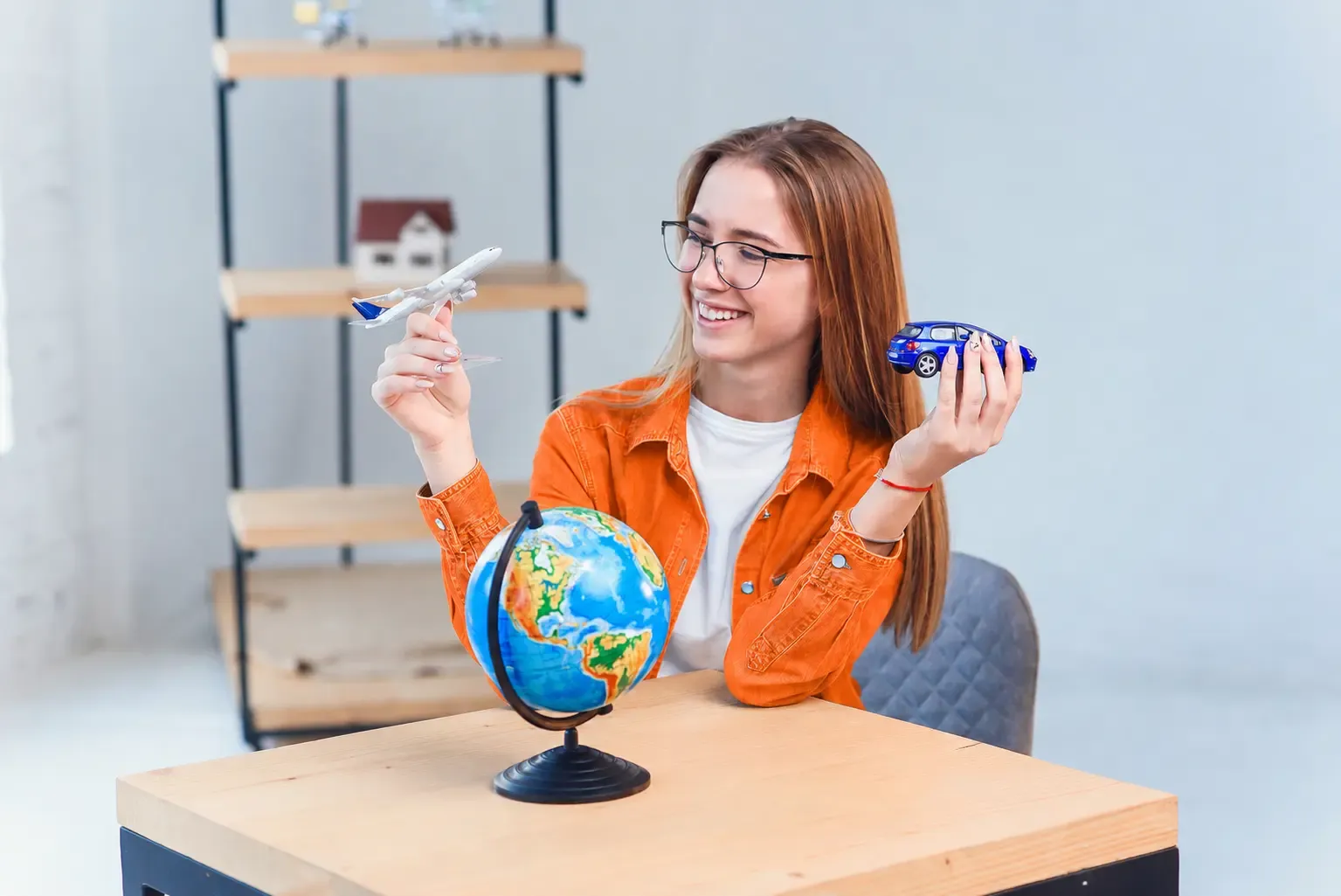 Young woman in an orange jacket sitting at a wooden table, smiling while holding a toy airplane and toy car beside a globe in a bright, travel-themed room.