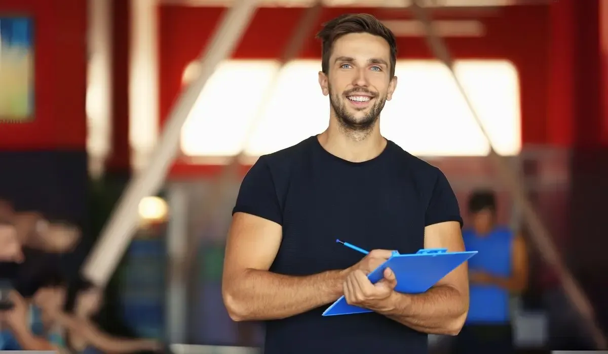 A friendly fitness trainer in a gym setting, holding a blue clipboard and smiling confidently, with a vibrant background of exercise equipment and people working out.
