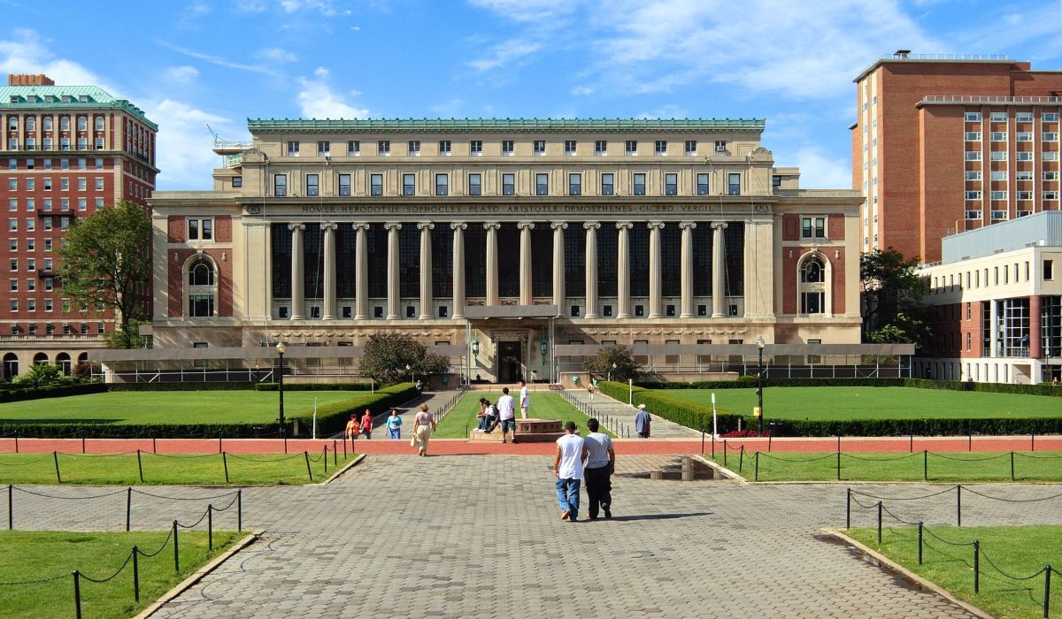 The grand neoclassical façade of Butler Library at Columbia University, with visitors walking along the tree-lined stone pathways on a clear day.