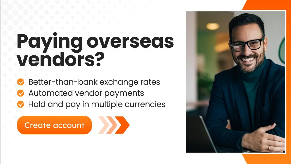 A professional man wearing glasses and a dark suit smiles while sitting at a desk with a laptop. The banner features text that says 'Paying overseas vendors?' and lists benefits such as better-than-bank exchange rates, automated vendor payments, and holding multiple currencies. A bright orange button reads 'Create account'.