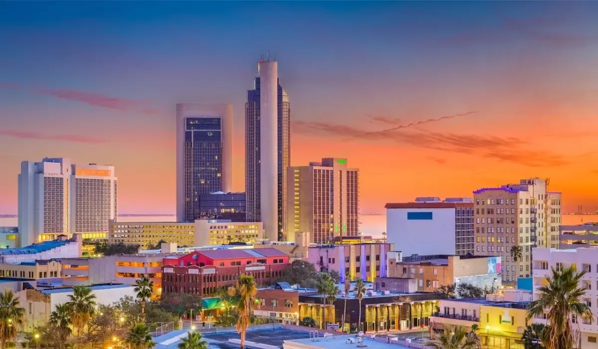 A scenic coastal view of Corpus Christi, Texas, featuring the marina with docked boats and the city skyline in the background under a clear blue sky.