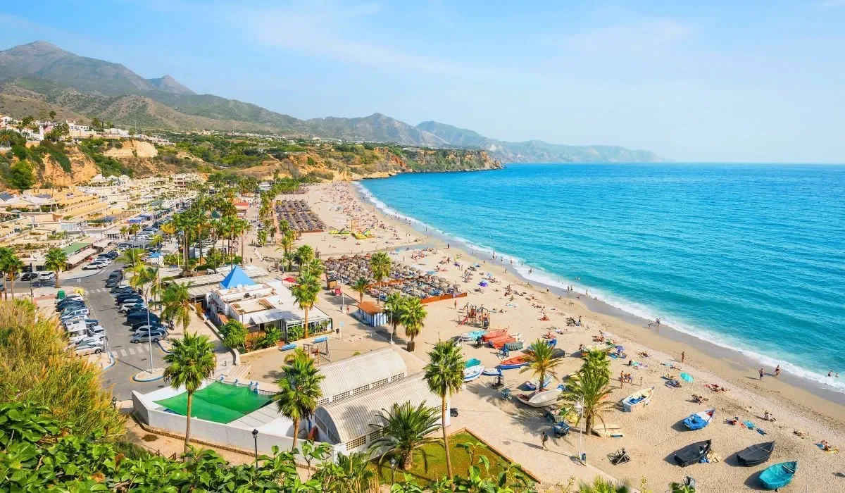 A scenic view of a sun-drenched beach lined with umbrellas and palm trees, backed by coastal mountains and a resort town in southern Spain.