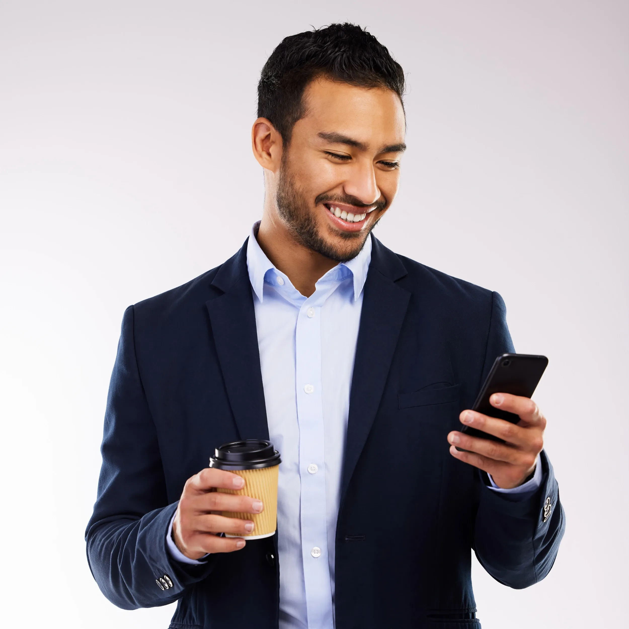 Smiling businessman in a suit holding a coffee cup and checking his phone, standing against a light background.
