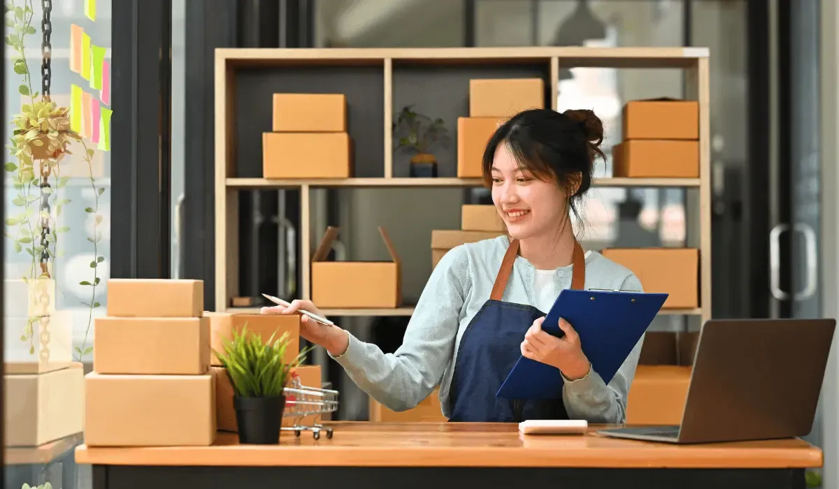 Ecommerce business owner organizing packages at a desk with a clipboard and laptop in a modern workspace.