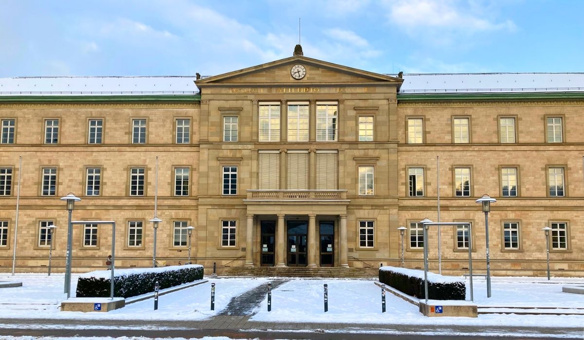 A winter scene at Eberhard Karls University of Tübingen, showing a symmetrical stone academic building with snow-covered grounds and a blue sky above.