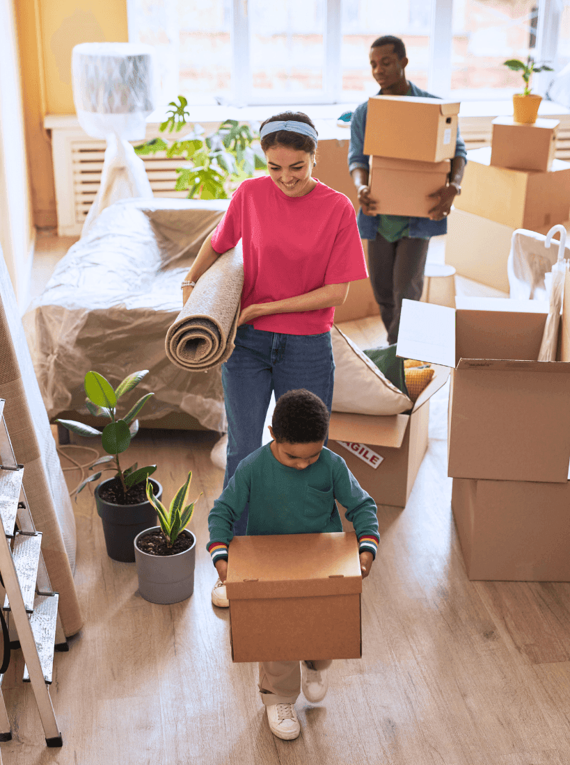 Family carrying boxes and a rug while moving into a new home surrounded by packed items