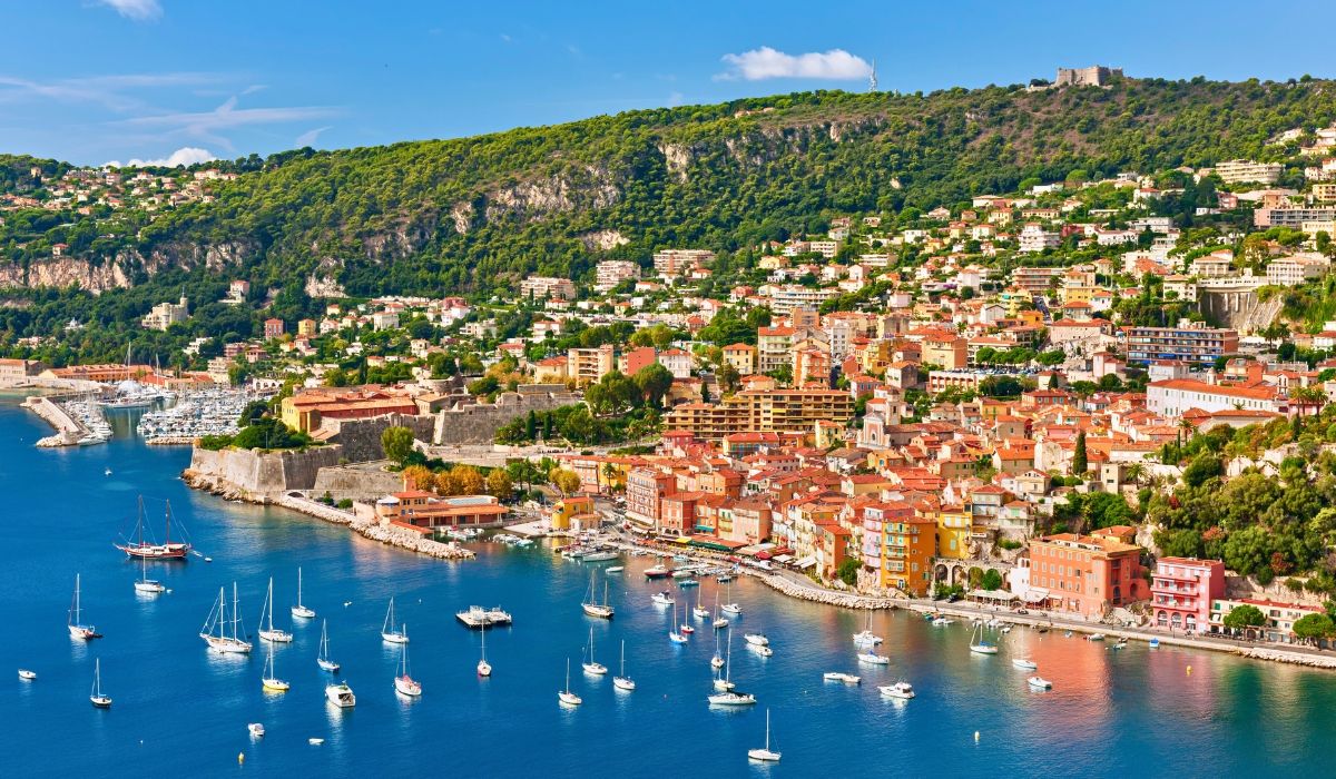 A picturesque coastal scene of the French Riviera, featuring turquoise waters, sandy beaches, and hillside villas under a bright sky.