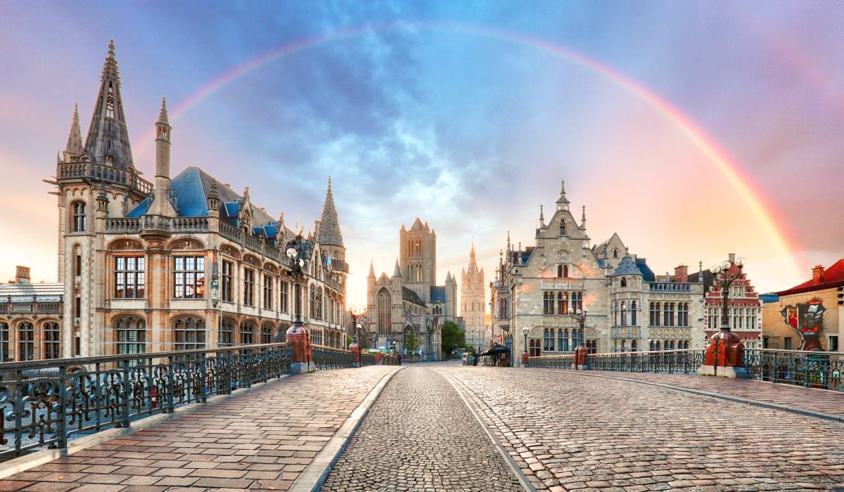 Picturesque medieval architecture of Ghent, Belgium, with a rainbow arching over the city’s iconic St. Nicholas’ Church and cobblestone bridge.