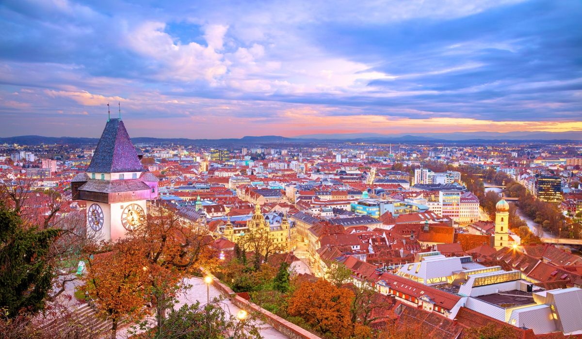 An aerial shot of Graz’s Old Town with red-tiled roofs, historic buildings, and the medieval clock tower (Uhrturm) surrounded by green hills.