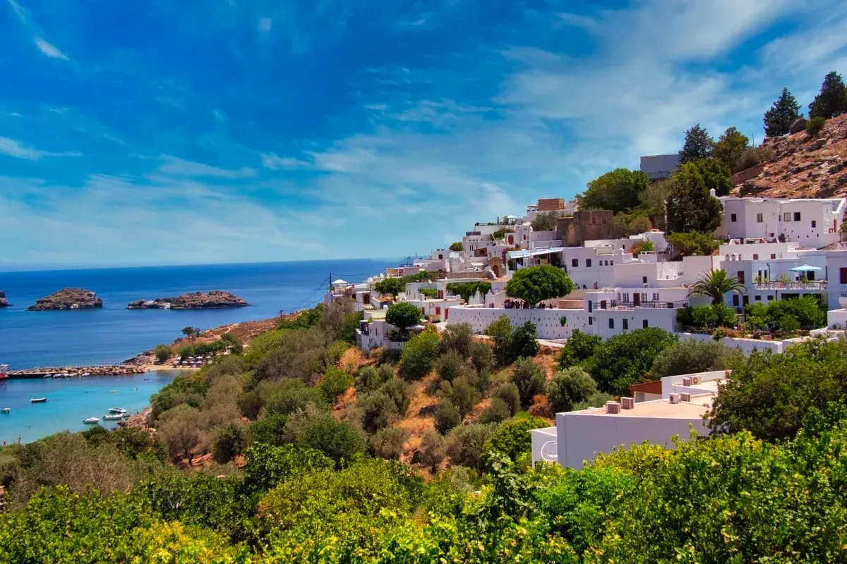 White hillside buildings overlook a turquoise bay and deep blue sea, surrounded by lush greenery under a sunny Mediterranean sky.