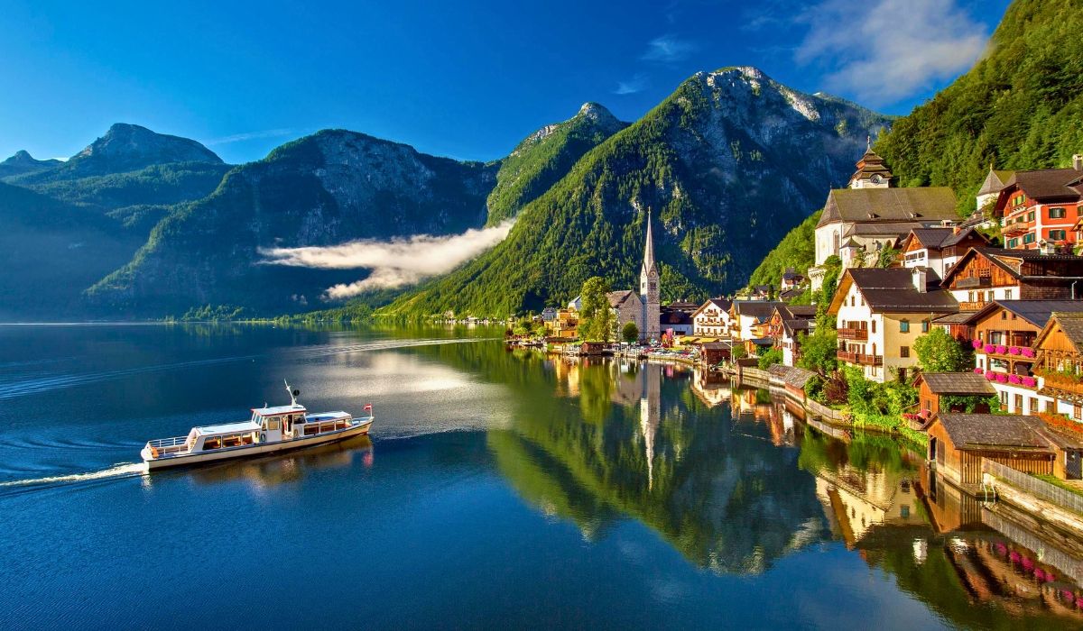 A tranquil lakeside scene of Hallstatt village with alpine-style homes, a church steeple, and mountains reflecting on calm waters.