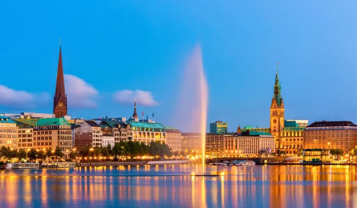 Evening view of Hamburg, Germany, with the illuminated Town Hall, church spires, and fountains reflecting on the Alster Lake. Canadians buying property in Germany can take advantage of competitive CAD to EUR exchange rates and low transfer fees through MTFX.