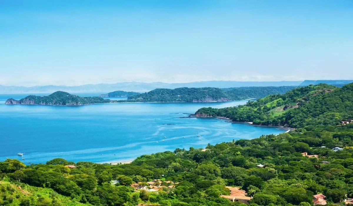 Aerial view of a lush green coastline and calm blue bay in Costa Rica, with scattered houses nestled among tropical hills.