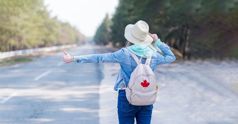Person with a backpack featuring a Canadian flag hitchhiking on an empty rural road, symbolizing travel or relocation abroad.