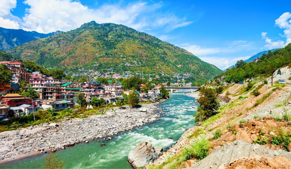 Mountain town in Himachal Pradesh, India, along a river with a scenic backdrop of green hills.