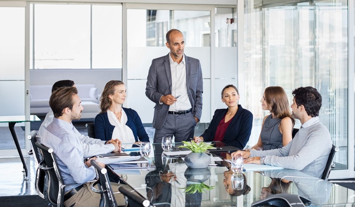Business professionals in formal attire engaged in a meeting around a conference table, with one man standing and presenting to the group in a modern office setting.