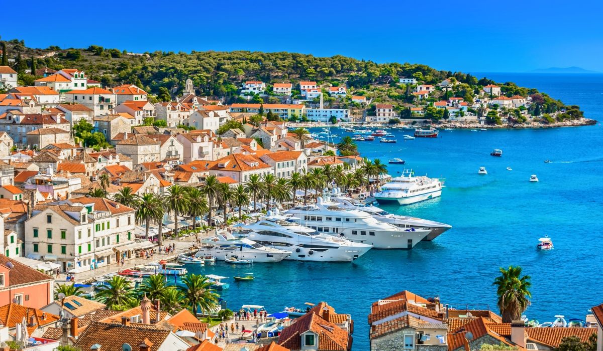 Picturesque harbor in Hvar filled with luxury yachts and stone houses, framed by palm trees and a green hillside.