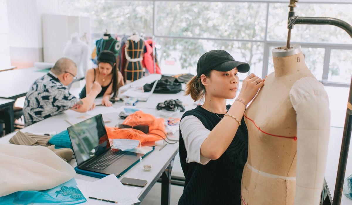 Hero image showing a fashion company office with a woman wearing a cap and fixing something on the dress on a mannequin and a few coworkers talking in the background.