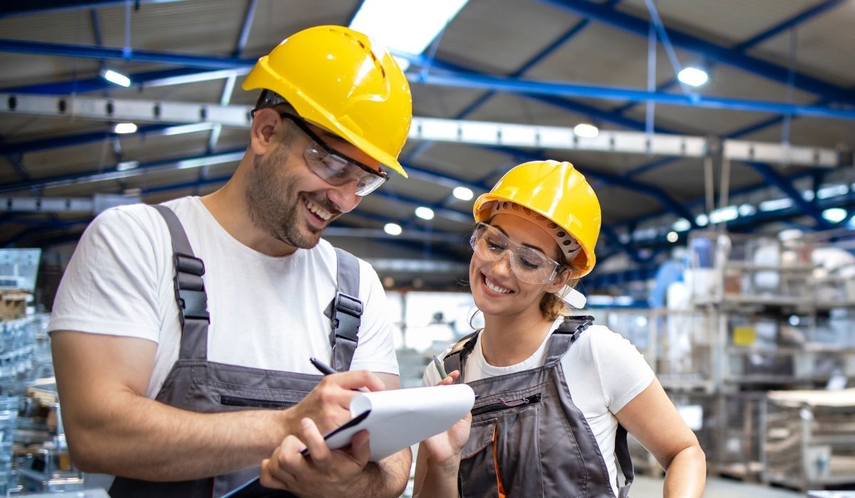 Safety hat wearing young male and female warehouse workers looking at the notepad with smiles on their faces.