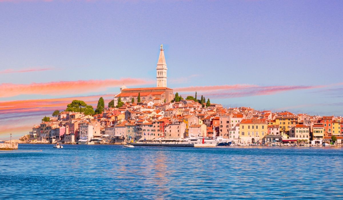 Coastal townscape of Rovinj in the Istrian Peninsula, featuring the Church of St. Euphemia and colorful buildings along the Adriatic under a pastel sunset sky.