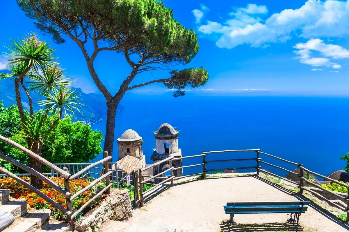Scenic coastal viewpoint with a pine tree, flowering garden, wooden railings, and historic stone towers overlooking the bright blue sea under a sunny sky.