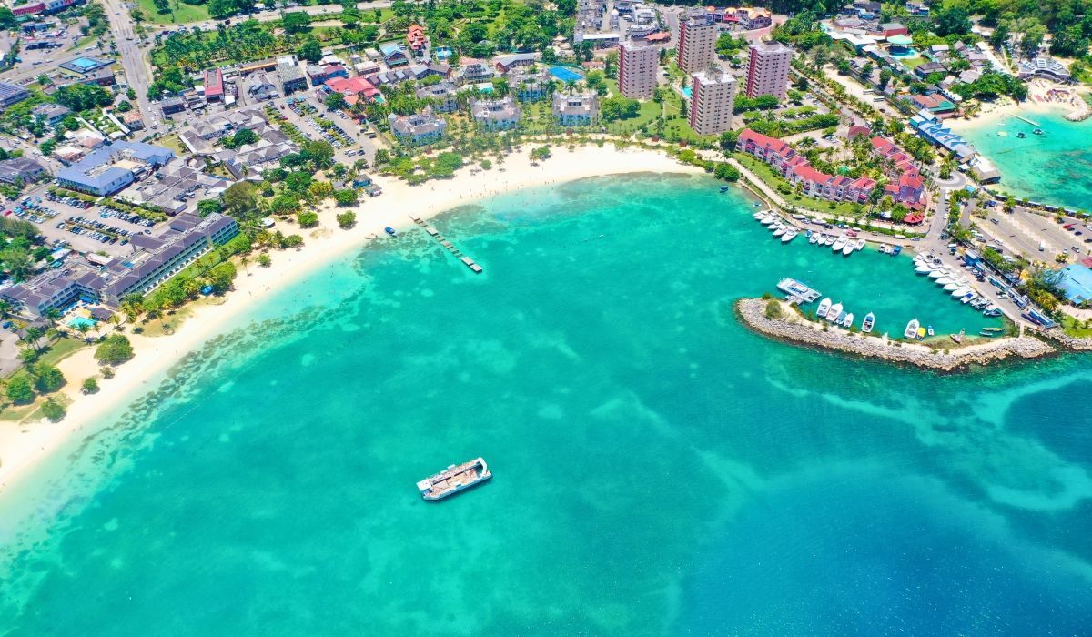 Aerial view of a turquoise bay in Jamaica with a docked boat, sandy beach, and coastal hotels and buildings.