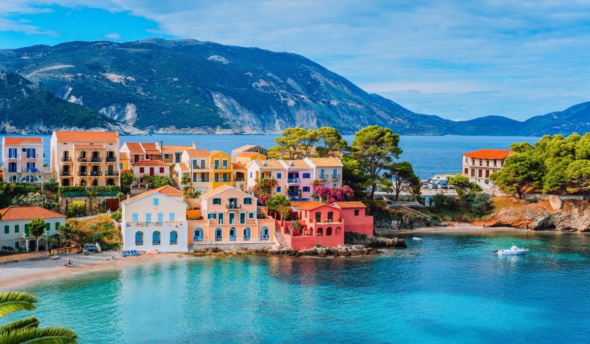 Colorful seaside village houses in Assos, Greece, with turquoise waters and mountainous terrain in the background.