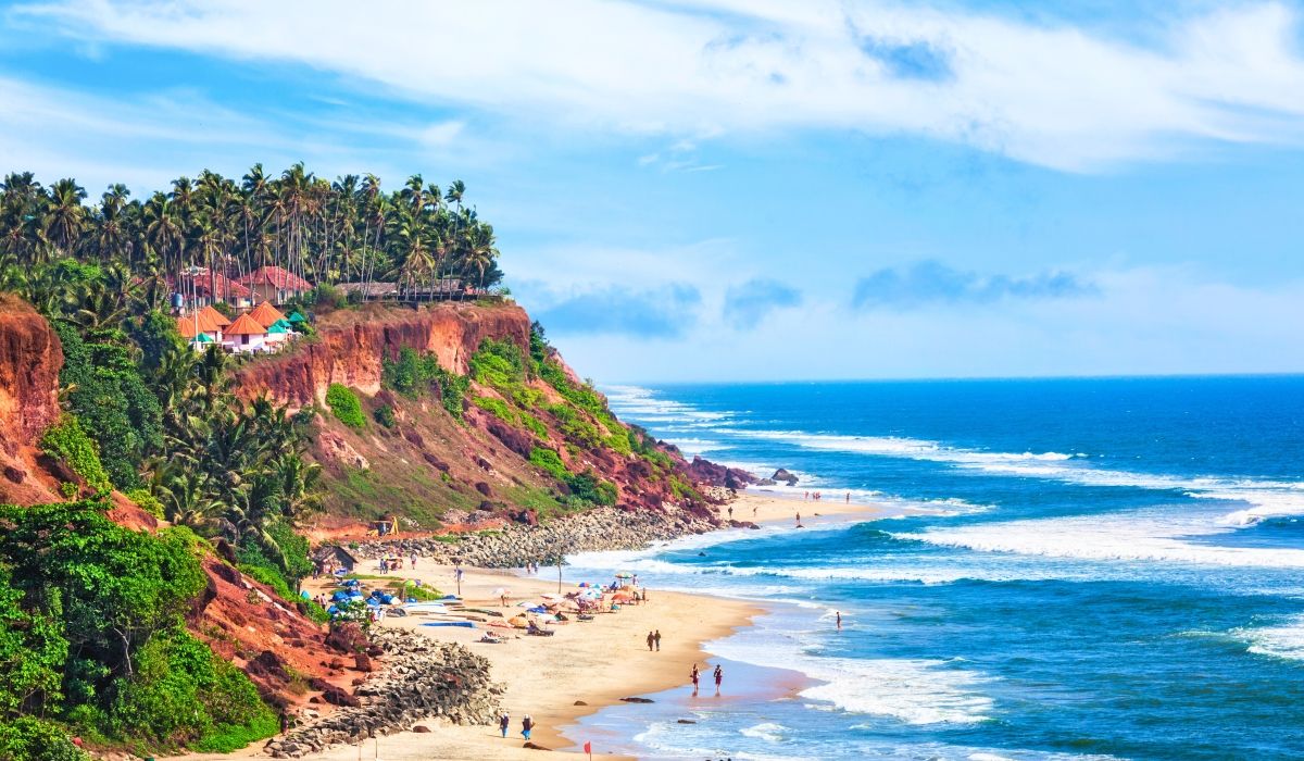 Scenic view of a tropical beach with red cliffs, palm trees, and beachgoers along the shoreline in Varkala, Kerala, India.