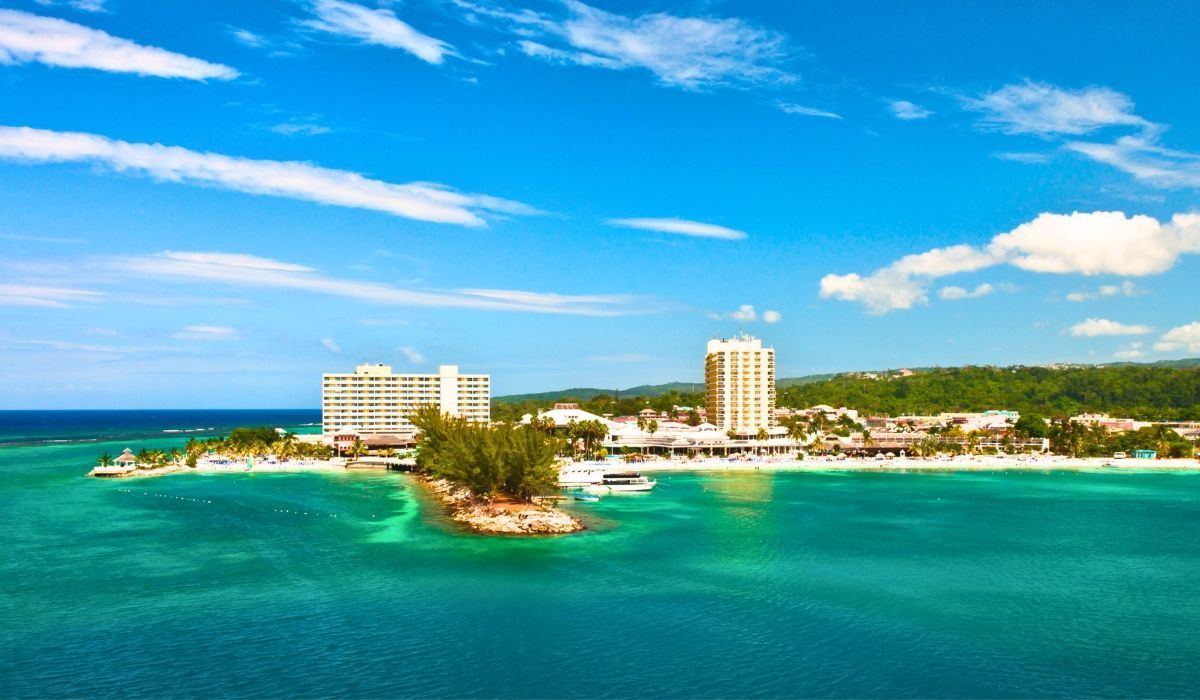 Scenic waterfront view of Kingston, Jamaica, featuring coastal resorts, high-rise buildings, and turquoise waters under a sunny blue sky.