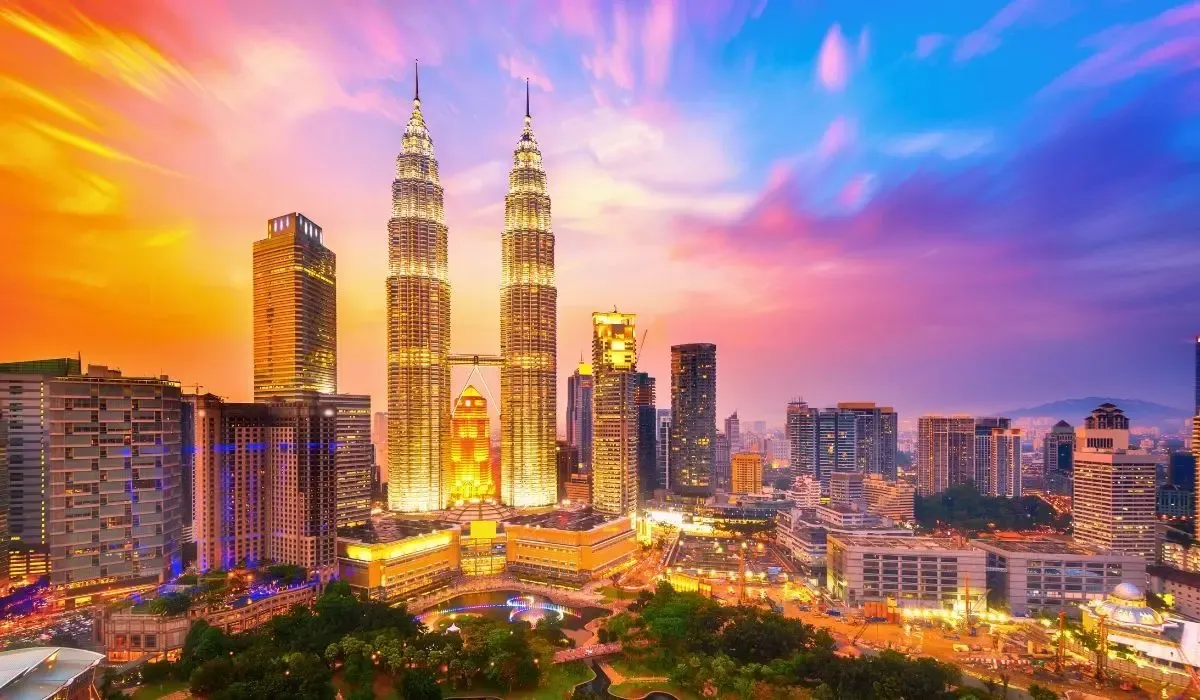 A cityscape of Kuala Lumpur, showcasing the iconic Petronas Twin Towers, modern high-rise buildings, and surrounding green spaces under a clear sky.