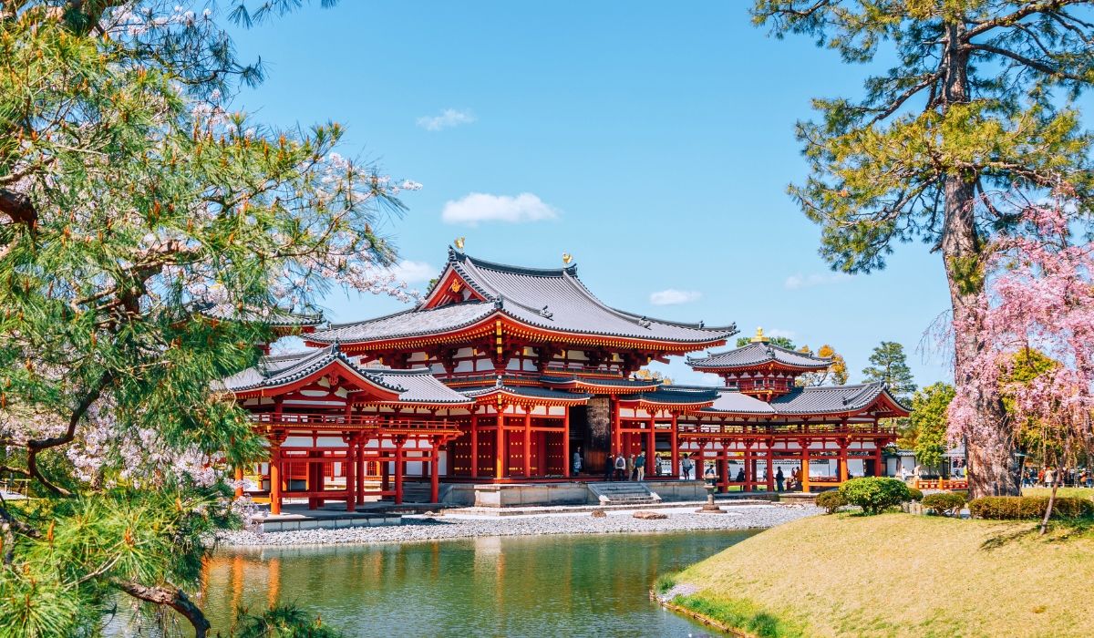 Historic Byodoin Temple in Kyoto framed by cherry blossoms and pine trees, with its reflection visible in a calm pond.