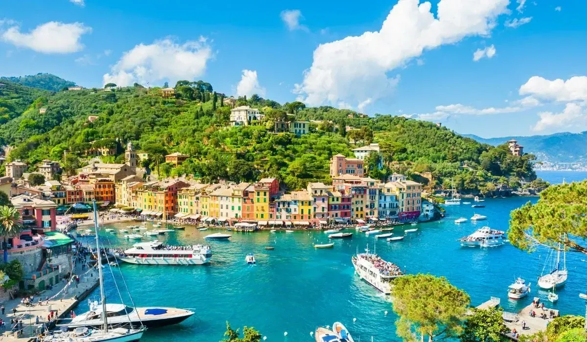 Colorful hillside village overlooking a turquoise harbor with boats and lush green mountains.