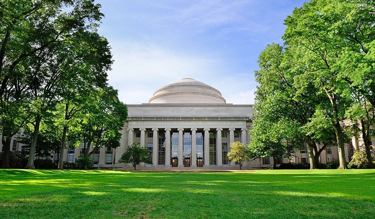 MIT’s iconic Great Dome surrounded by leafy green trees and a wide open lawn, bathed in bright springtime sunlight.