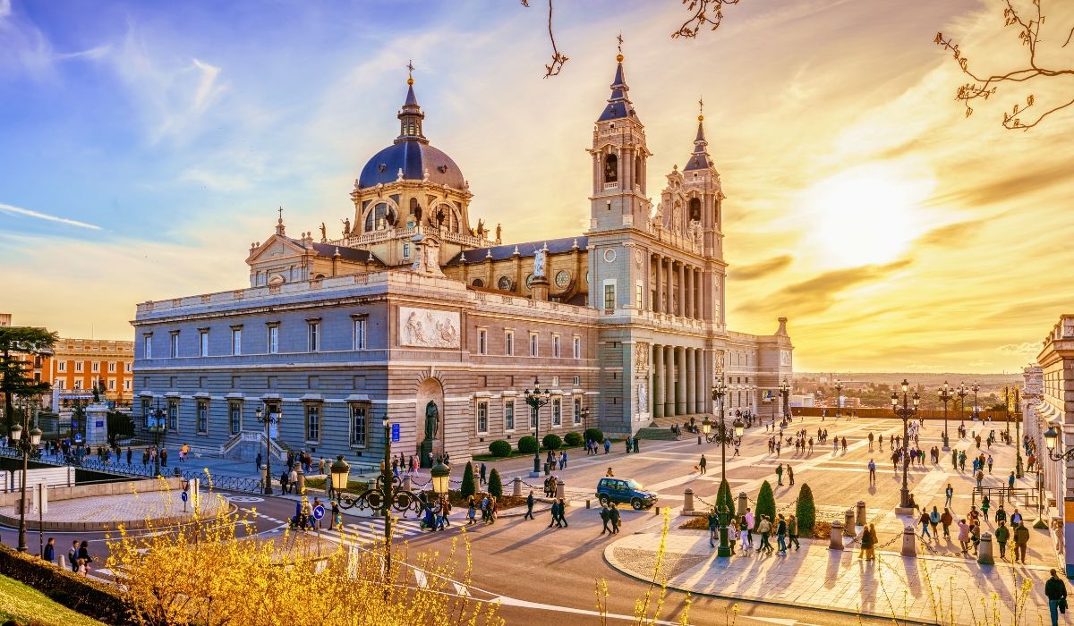 A golden sunset illuminating Almudena Cathedral in Madrid, with its neoclassical and Gothic facade, twin towers, and a large square filled with pedestrians.