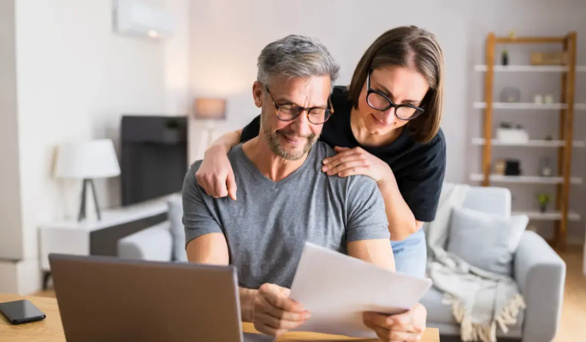 A middle-aged couple with glasses sitting at a table, reviewing financial documents together. The man wears a gray t-shirt and holds the papers, while the woman, wearing a black shirt, leans over his shoulder smiling. A laptop and smartphone are on the table in a cozy, well-lit living room.