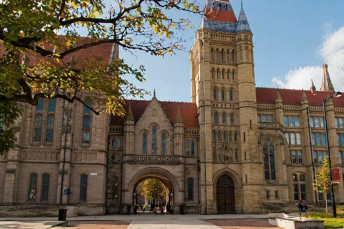 Historic Gothic-style entrance of the University of Manchester with sandstone architecture, arched gateway, tall tower, and trees framing the campus scene.