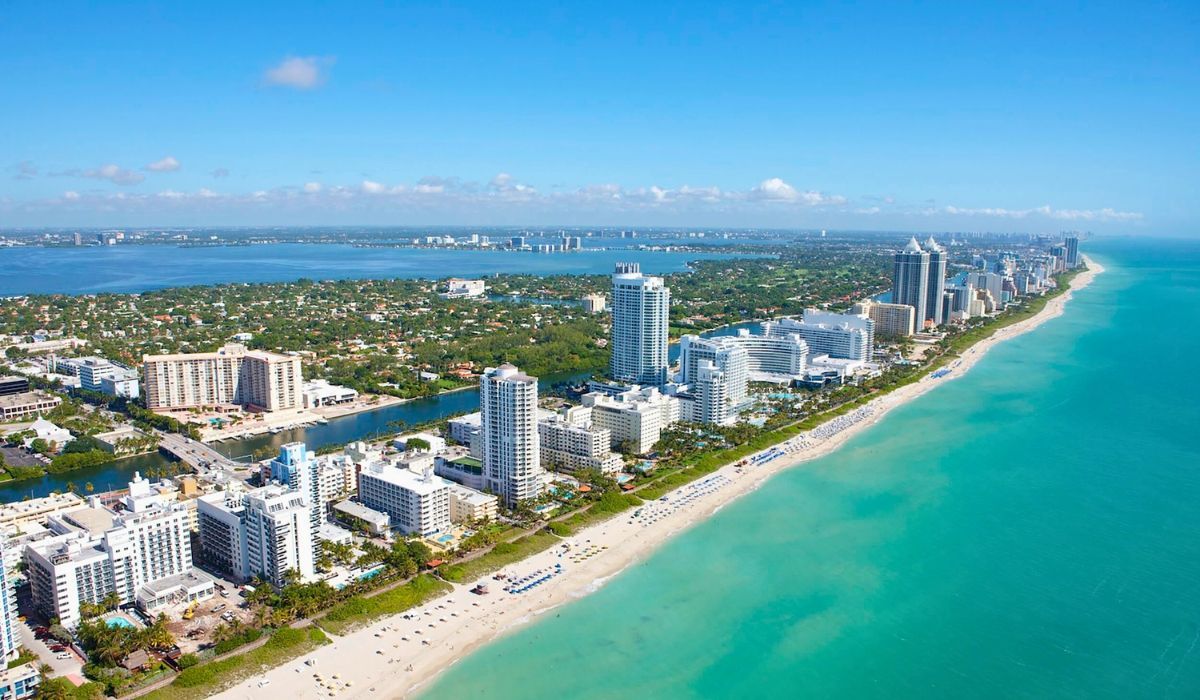 A vibrant scene of Miami Beach, Florida, featuring Art Deco architecture along Ocean Drive, palm-lined streets, and the Atlantic Ocean's blue waters under a bright sky.