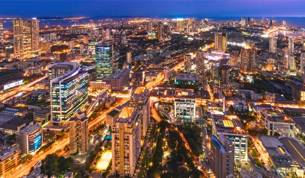 Aerial night view of Mumbai's cityscape with illuminated buildings, busy roads, and distant coastline.