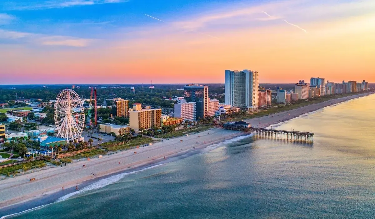A beachfront scene in Myrtle Beach, South Carolina, featuring a wide sandy shore, ocean waves, and a bustling boardwalk with attractions.