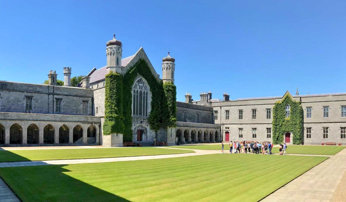 An aerial view of the National University of Ireland, Galway campus, featuring historic stone buildings surrounded by lush greenery and the River Corrib flowing nearby.