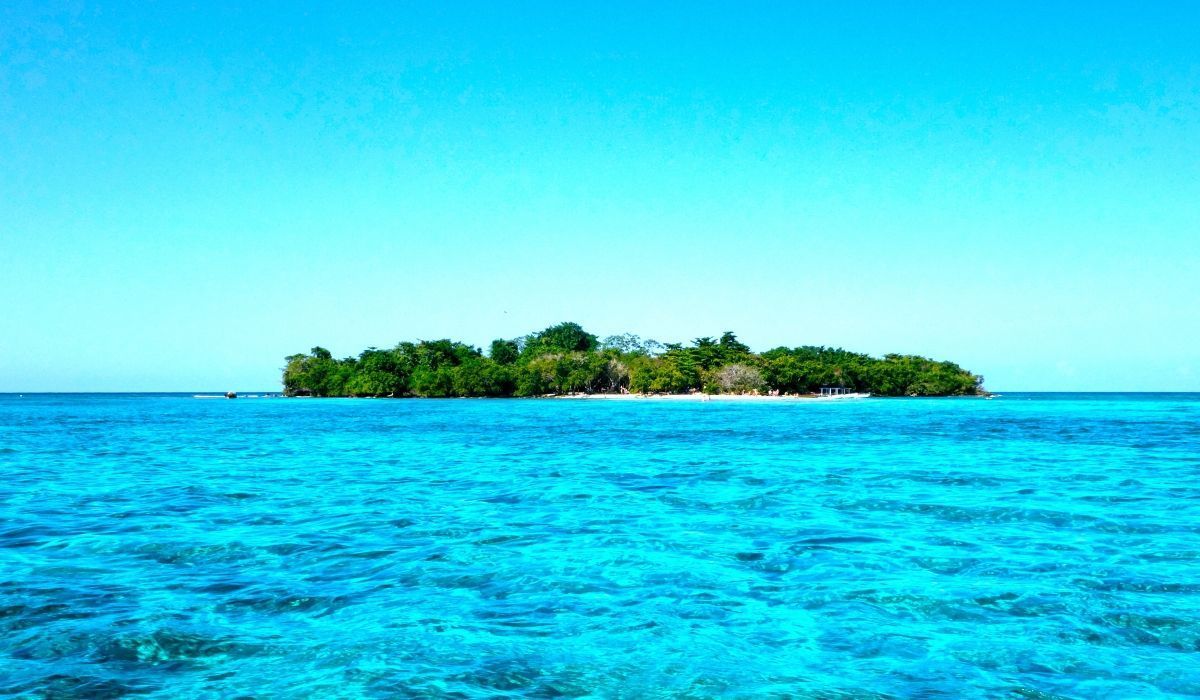 View of a small, lush island off the coast of Negril, Jamaica, surrounded by clear turquoise waters under a bright blue sky.