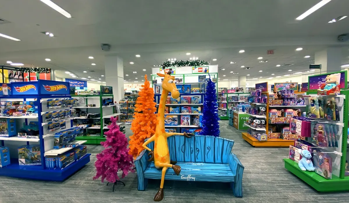 Colorful toy store display featuring Geoffrey the Giraffe on a bench surrounded by vibrant Christmas trees and popular toy brands like Hot Wheels and LEGO