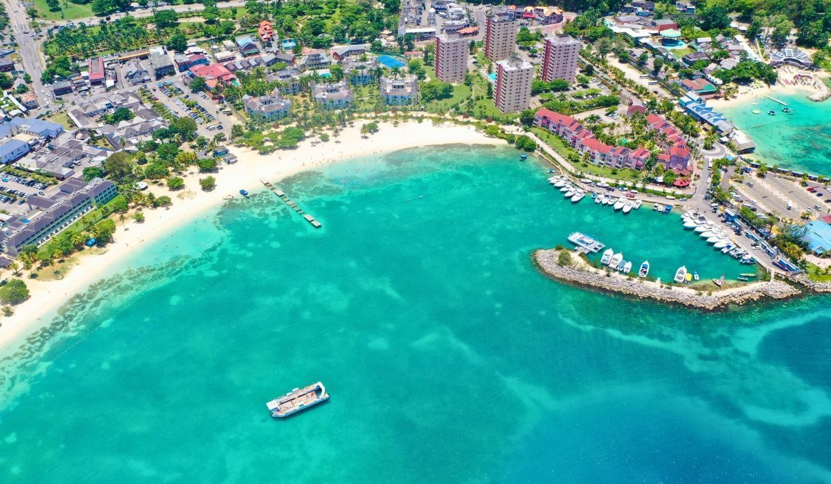 Aerial view of Ocho Rios, Jamaica, featuring turquoise waters, a beachfront lined with hotels and boats, and surrounding tropical greenery.