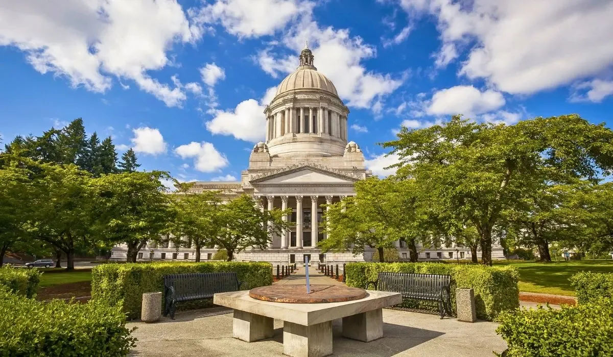 A scenic image of Olympia's waterfront featuring the Washington State Capitol building with its distinctive dome, set against a backdrop of evergreen trees and a clear blue sky.